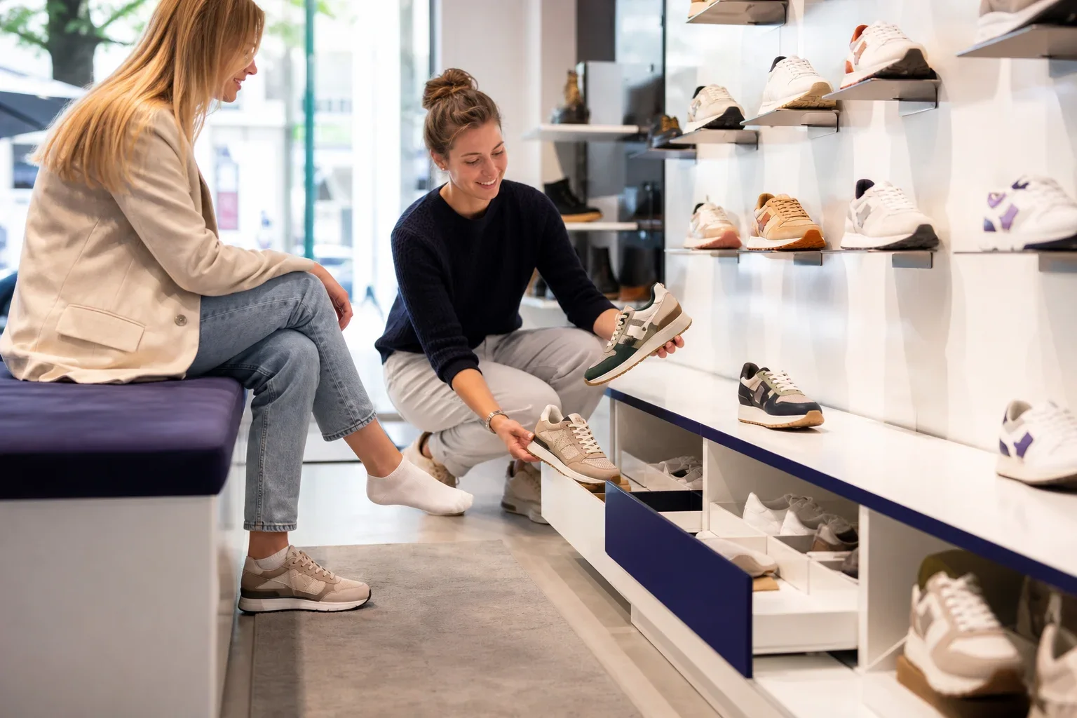 Waarom een Kassasysteem schoenenwinkel meer moet kunnen: Een winkeladviseur staat naast een zittende klant; beide personen zijn van opzij zichtbaar, met de schoenen en de pasplek als visueel middelpunt.; De adviseur haalt twee uitvoeringen